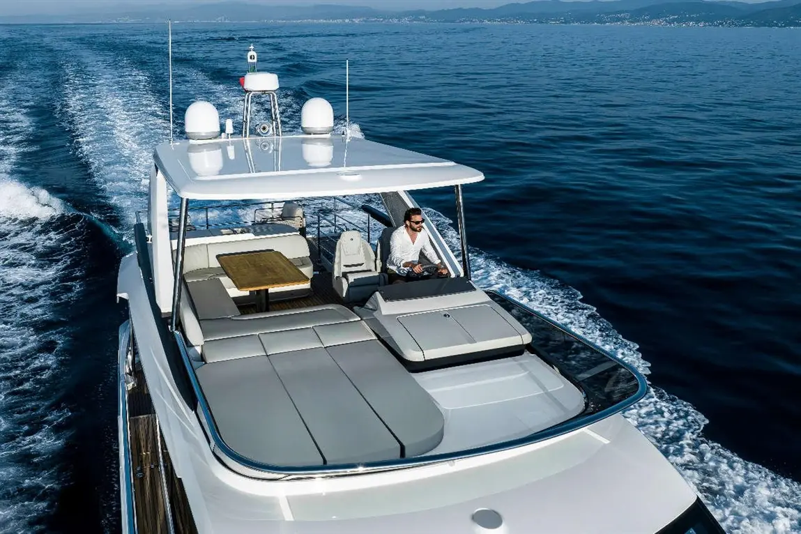 A man steers a luxury yacht through calm waters, featuring modern seating and a wooden table on the deck.